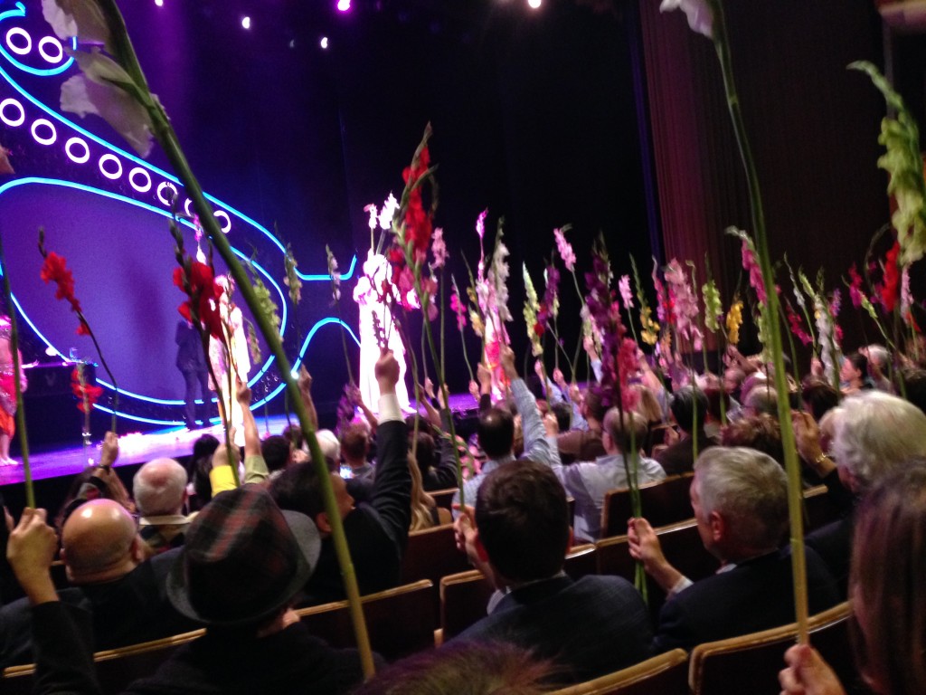 All of the orchestra-seated audience members waving their gladiolas along with Dame Edna, (who's lit-up all white on stage,) at her last opening night in LA, in 2015. Photo by Karen Salkin.